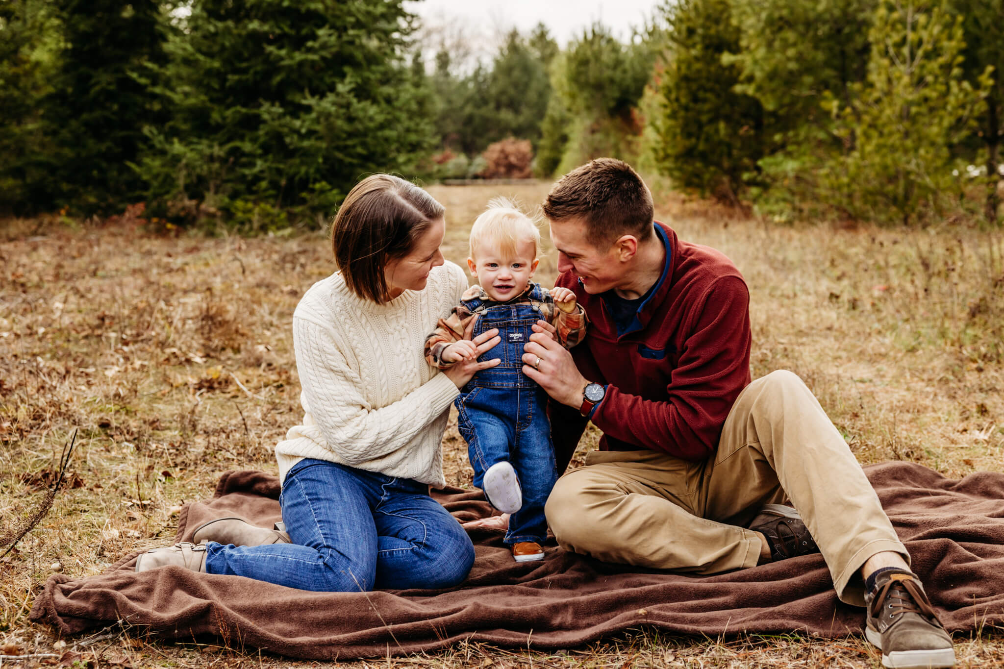 mom and dad tickling their little boy as they sit in a field of Christmas trees