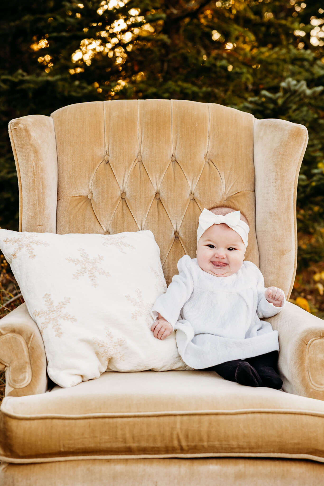 baby girl in a white outfit sitting on a tan velvet chair with a snowflake pillow for her Christmas photos