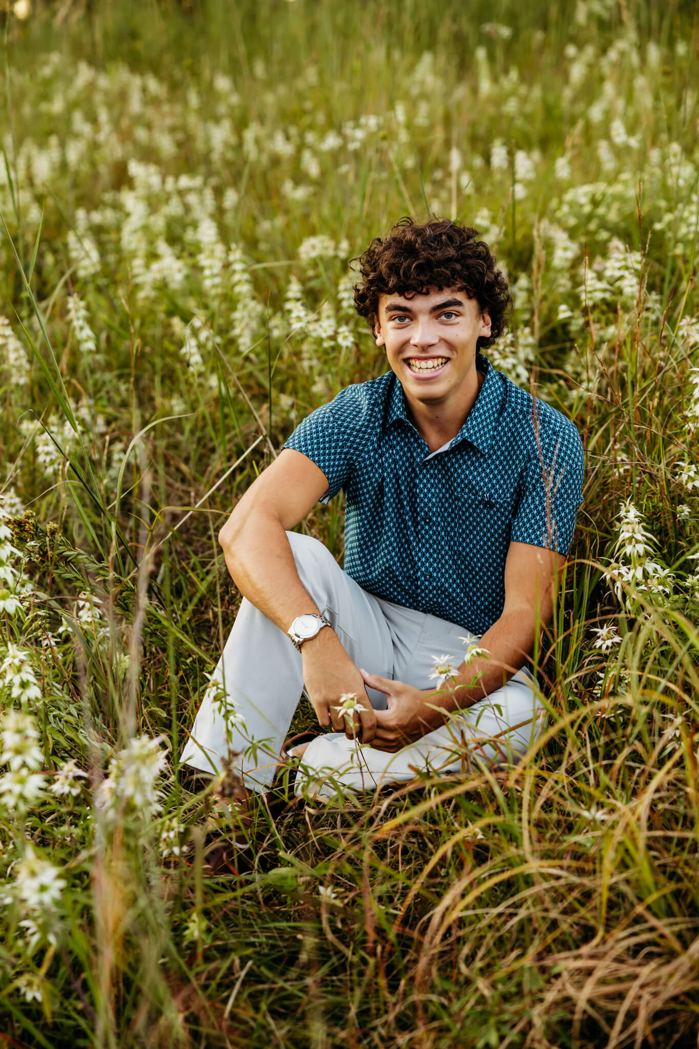 Teen boy laughing as he sits in the flowers at Tellulah Park for a blog about boys senior photos at Appleton East High School