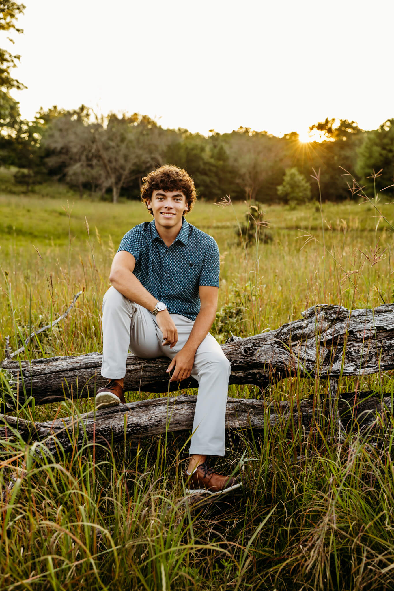 Appleton East High School senior boy sitting on a fallen tree and smiling as the sun sets behind him.