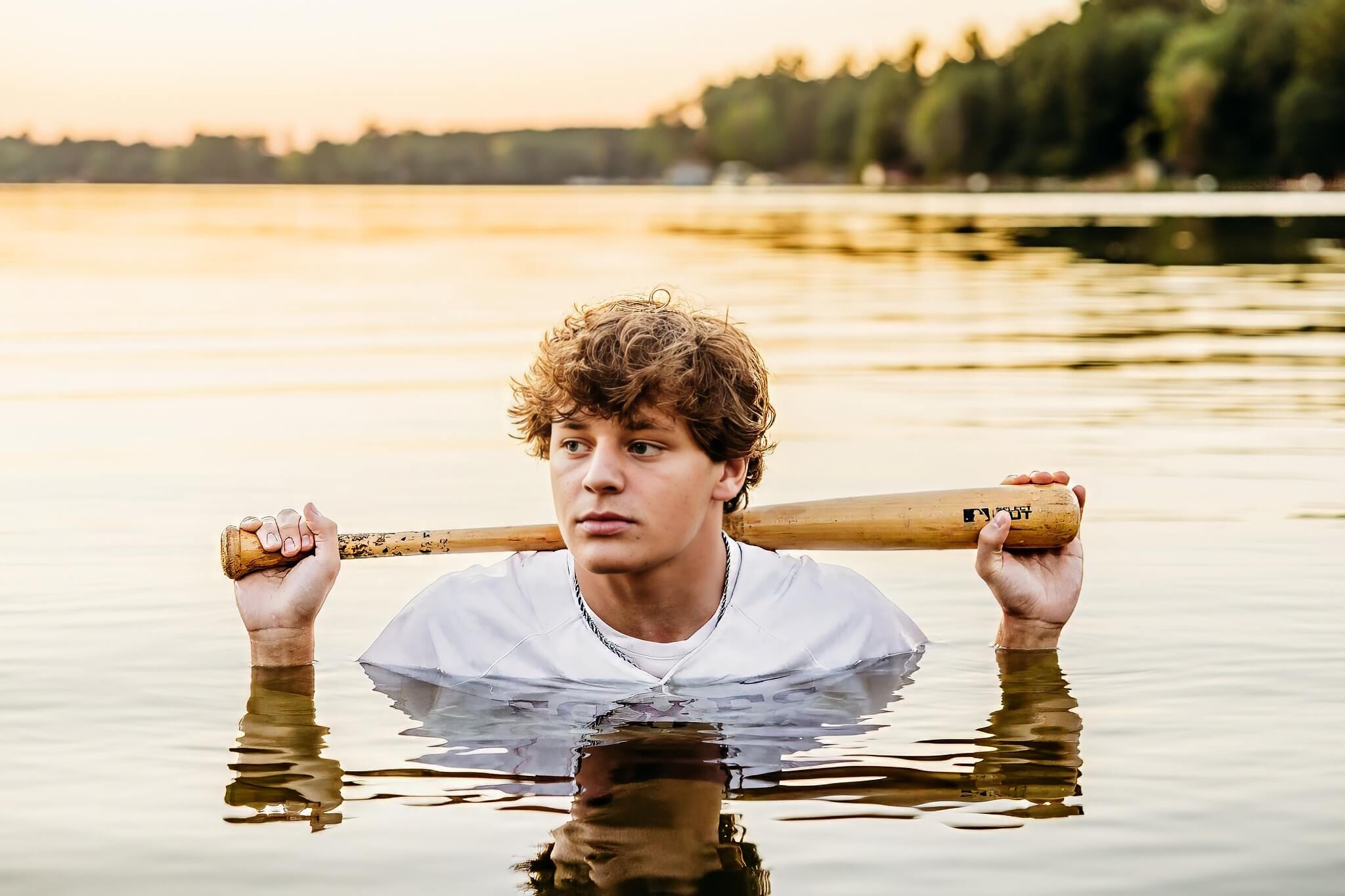 High school boy standing deep in the water as he rests his baseball bat on his shoulder at sunset.