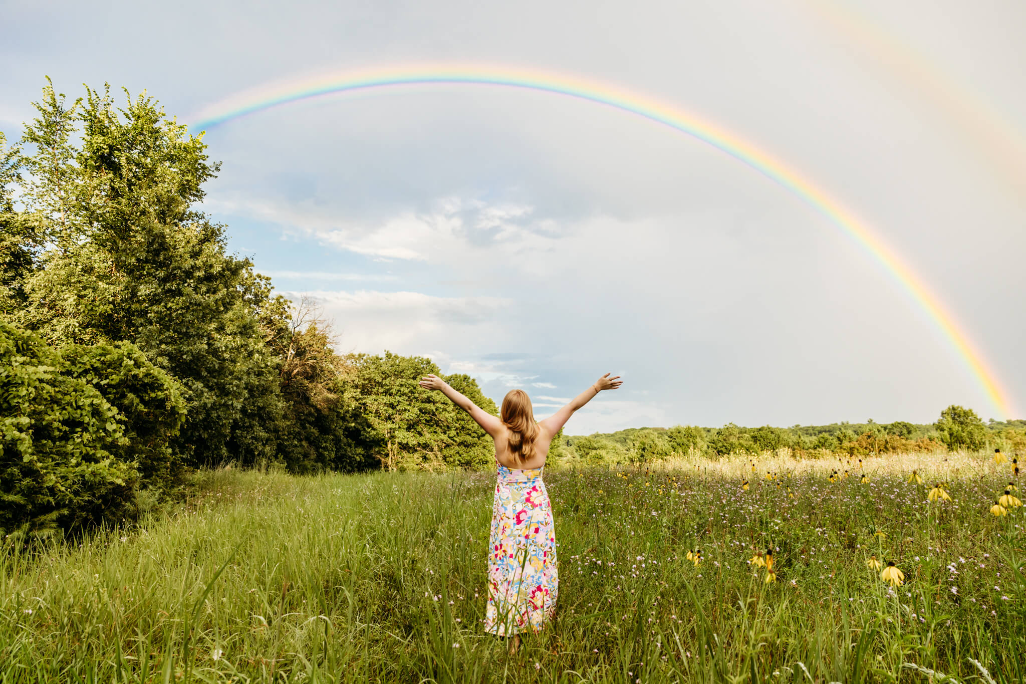 Beautiful young woman standing with her hands in the air and she looks up towards a rainbow in the sky.