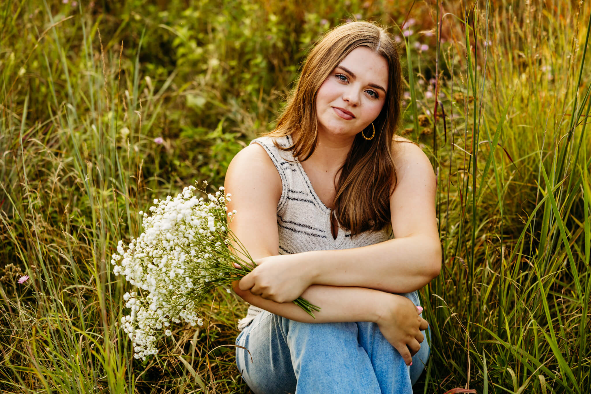 Young woman in a striped tank top and jeans holding a flower bouquet as she sits in tall grass for her senior photography session.
