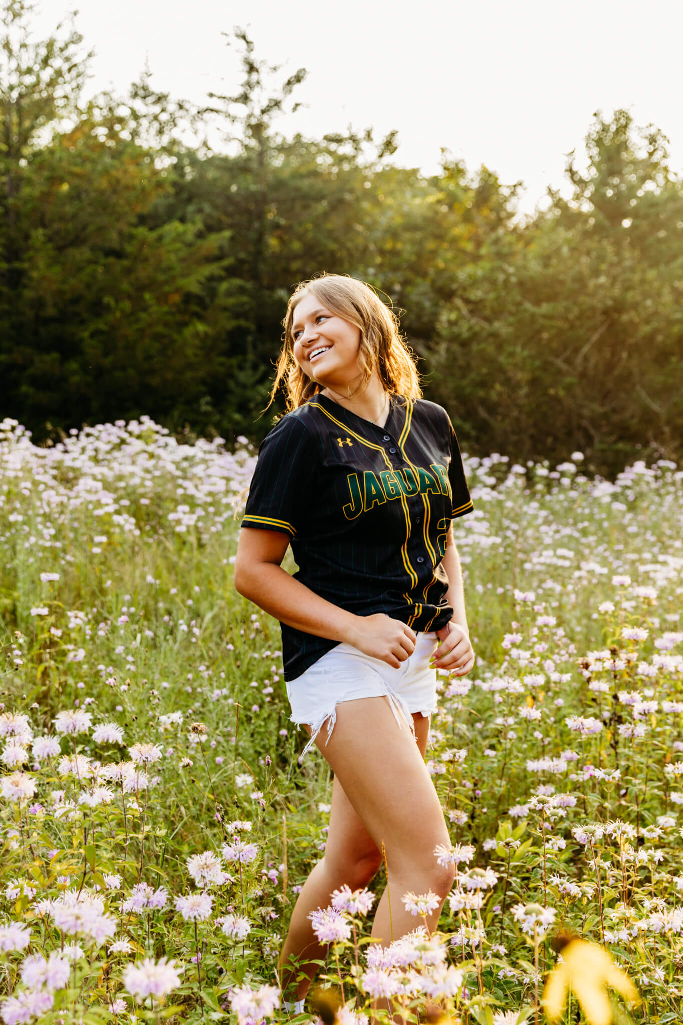Teen girl in a softball jersey and shorts smiling as she looks over her shoulder while standing in a wildflower field for her senior photo session.