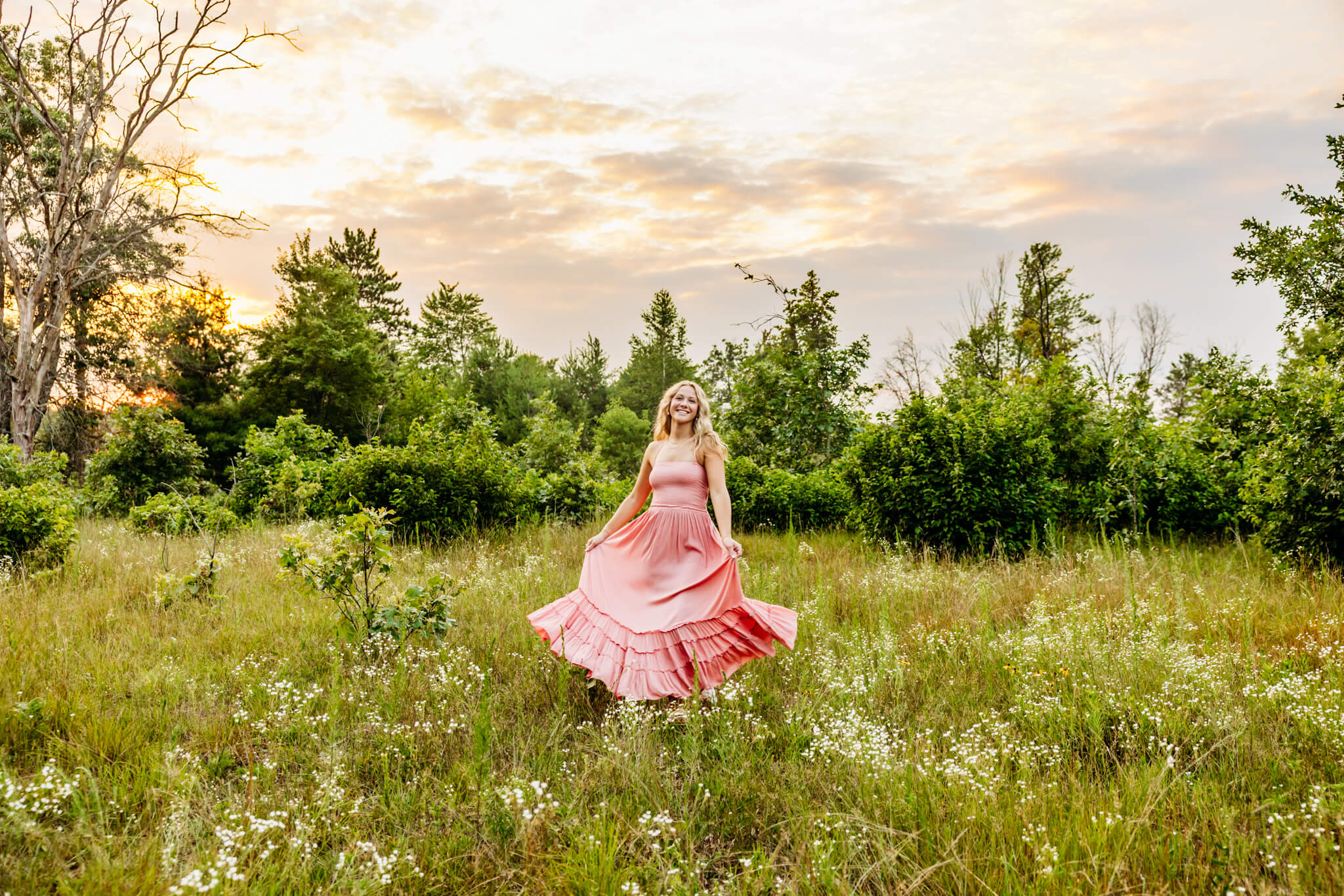 Young woman in a coral colored dress spinning and laughing in a wildflower field as the sun sets behind her.
