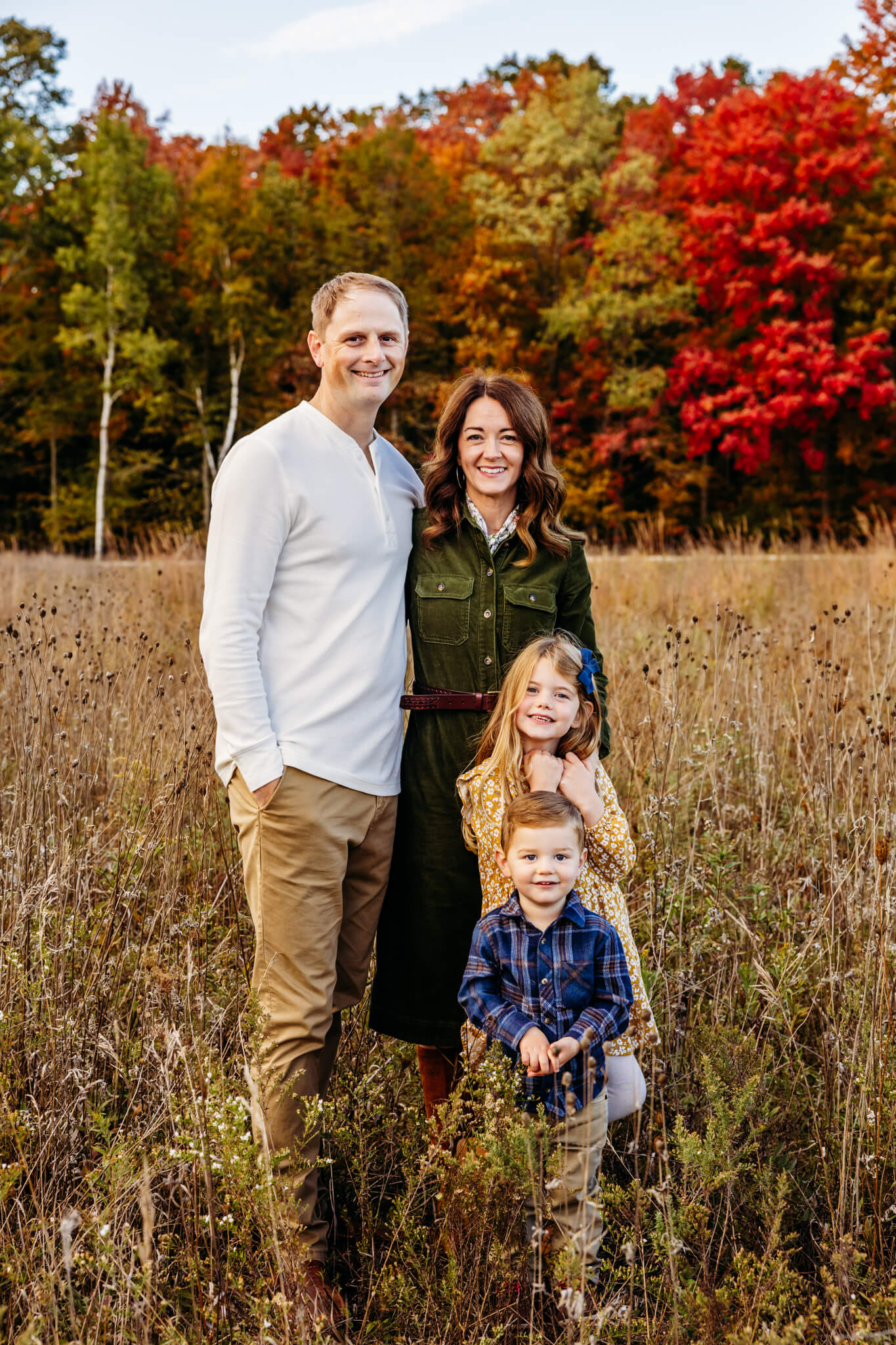 Beautiful family of four standing in a pumpkin patch with fall trees behind them as they enjoy their family photo session.